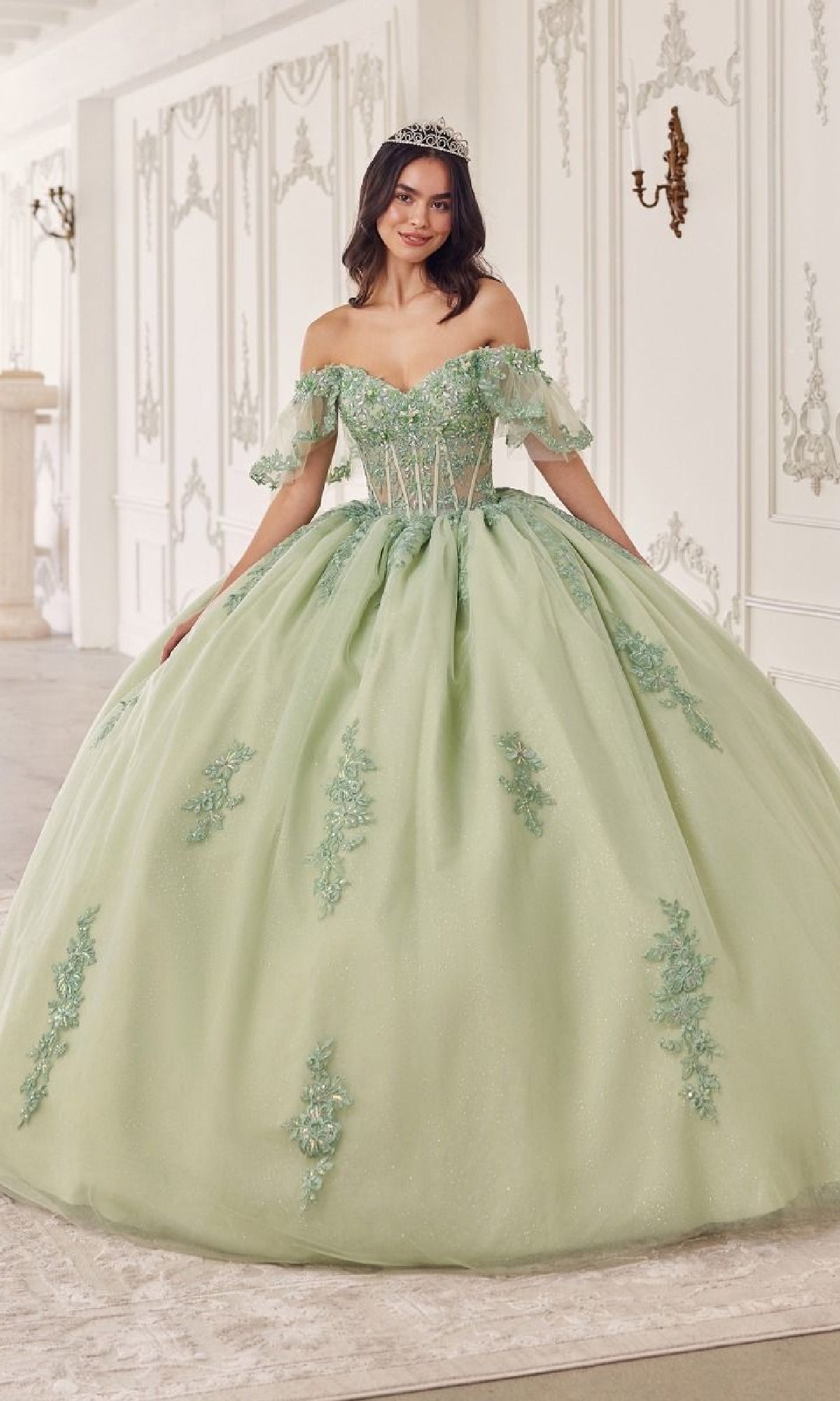 A young woman wearing Quinceanera Dress 15725 by Ladivine in sage green, with off-the-shoulder floral lace appliques and a tiara, stands gracefully in an ornate white room.